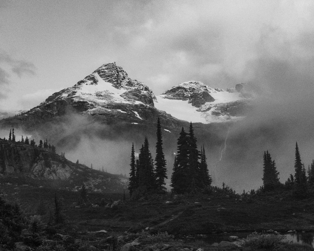 Black-and-white photograph of snow-dusted peaks and dark evergreens surrounded by mist.
