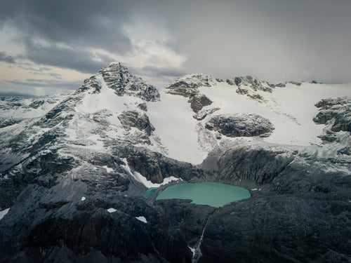 Snow-covered mountains surround a turquoise alpine lake beneath gray clouds.
