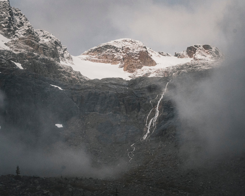 A narrow waterfall descends a dark mountain face below snowy peaks and drifting mist.