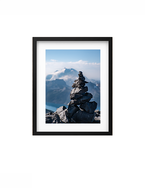 A framed landscape photograph of a mountain peak with a balanced rock cairn in the foreground, shown against a clean white background.