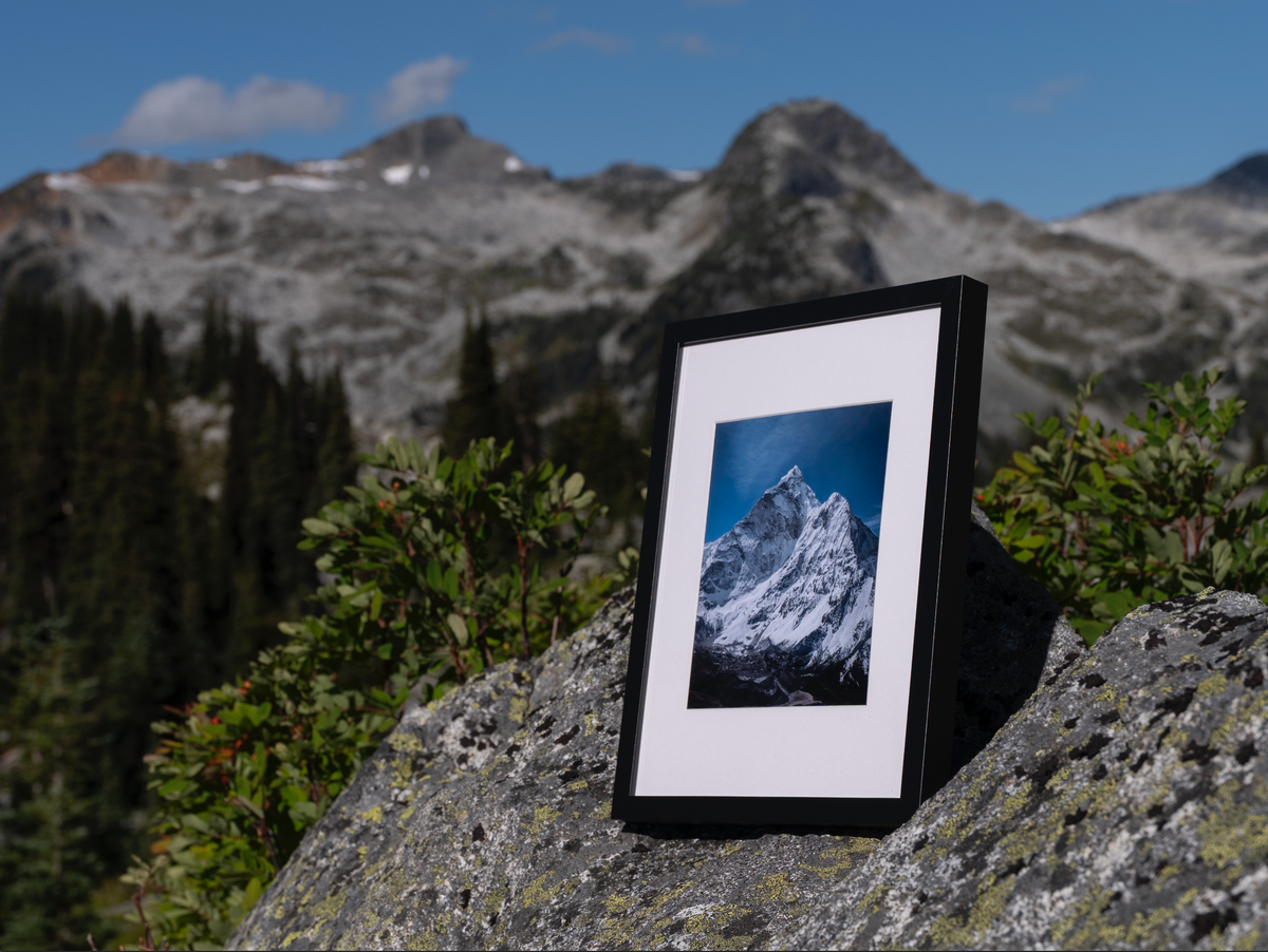 Solitude print in a black frame resting on a rock with mountains and trees behind it.